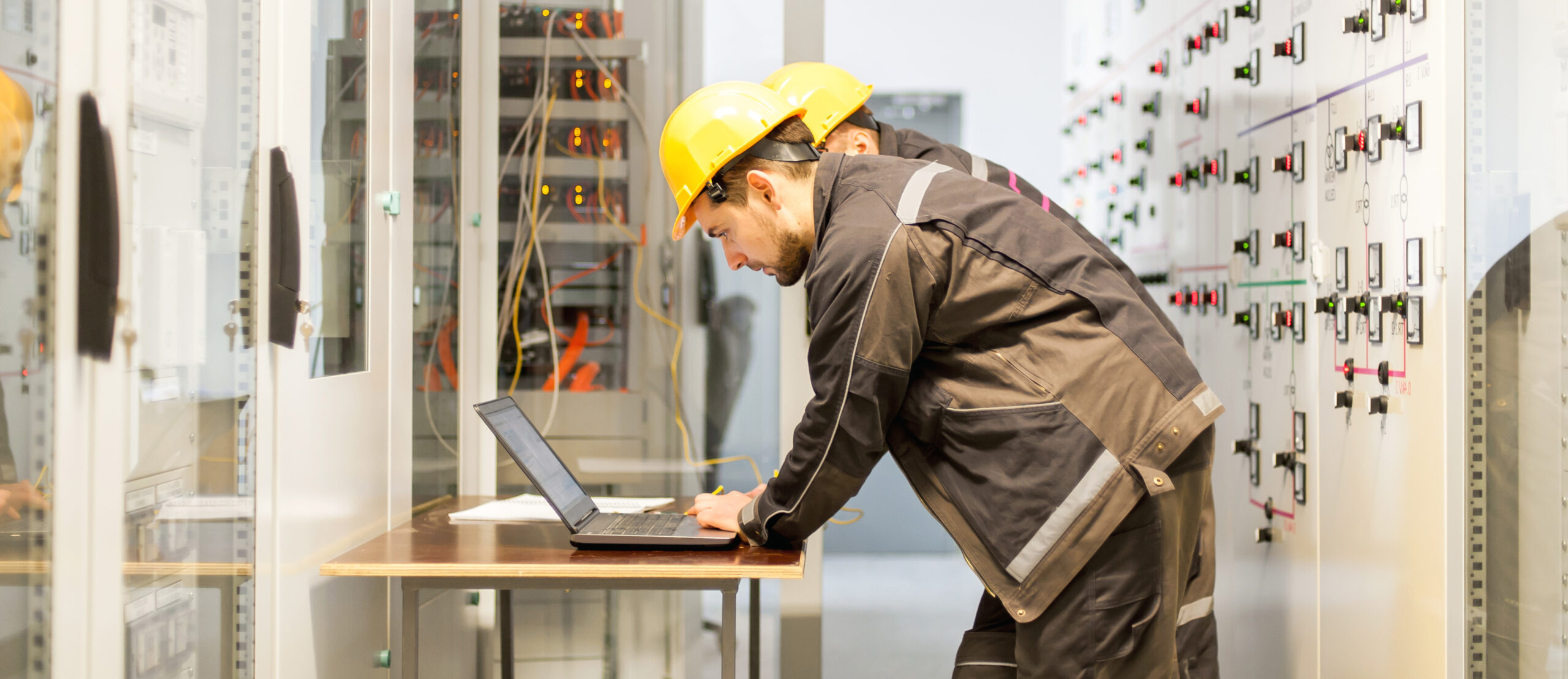 Two maintenance engineers inspect relay protection system with laptop computer. Bay control unit. Medium voltage switchgear